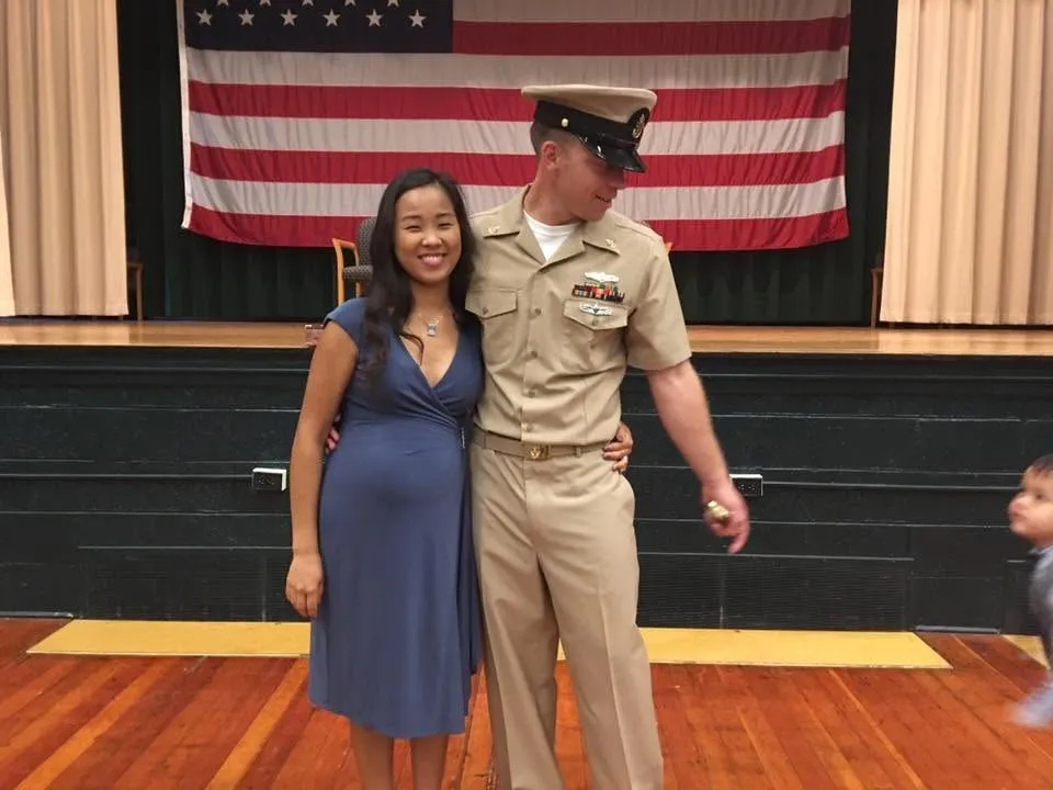 Oliver in khaki chief's uniform and cover, his arm around his wife, on a stage with a large American flag behind them.