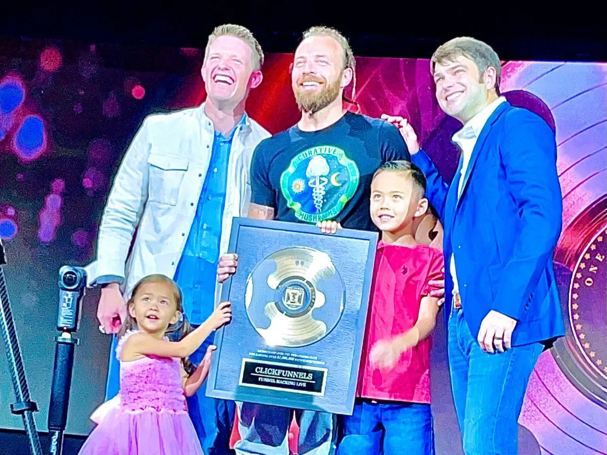 Oliver Carlin on stage receiving the ClickFunnels Two Comma Club award, with his children beside him.